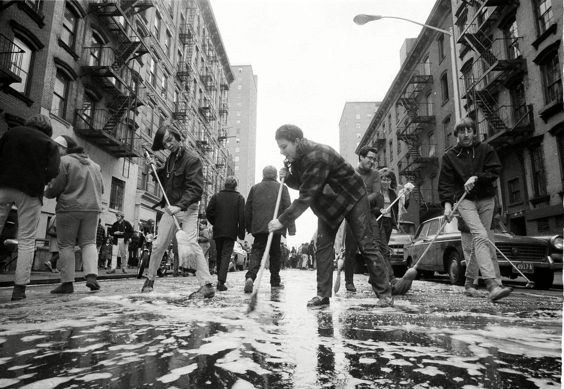 The caption reads: "East Village youths sling mops and brooms in the middle of the street during a neighborhood clean-up on East 3rd Street." April 1967.